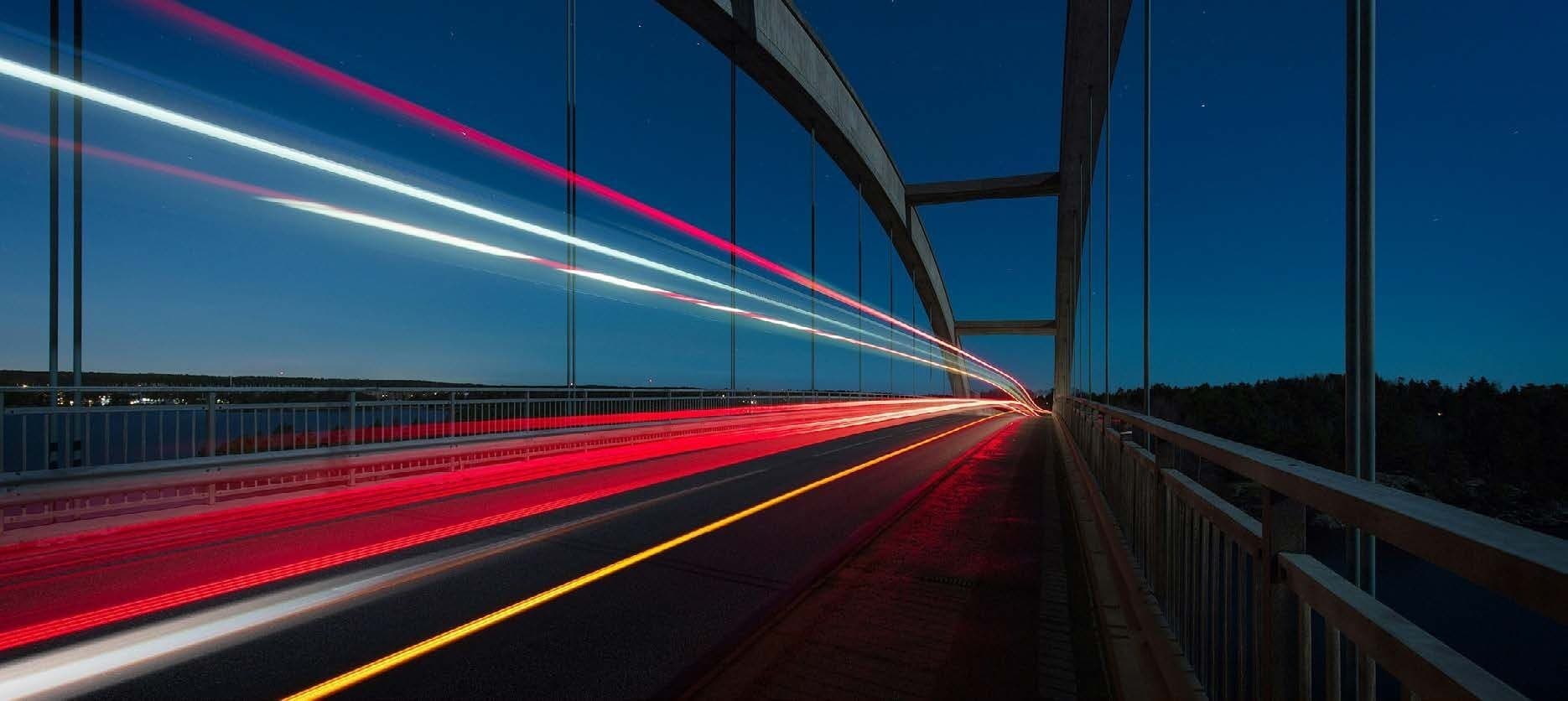 Long-exposure photo of a road bridge at dusk, with red, white and amber light trails streaming towards the far end under a deep blue sky
